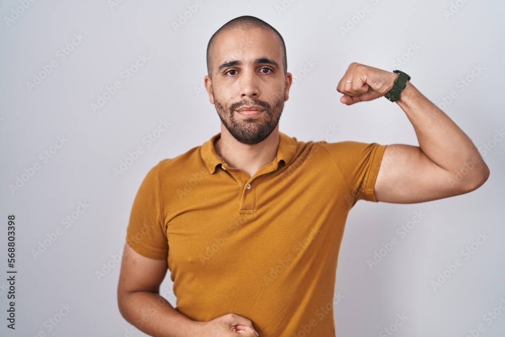 Hispanic man with beard standing over white background strong person showing arm muscle, confident and proud of power Hispanic man with beard standing over white background strong person showing arm muscle, confident and proud of power