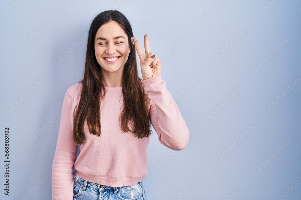 Young brunette woman standing over blue background showing and pointing up with fingers number two while smiling confident and happy. Young brunette woman standing over blue background showing and pointing up with fingers number two while smiling confident and happy.