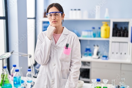 Young brunette woman working at scientist laboratory thinking concentrated about doubt with finger on chin and looking up wondering