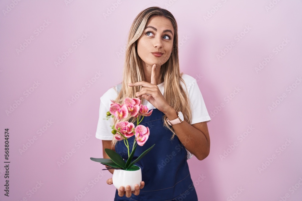 Young blonde woman wearing gardener apron holding plant thinking concentrated about doubt with finger on chin and looking up wondering