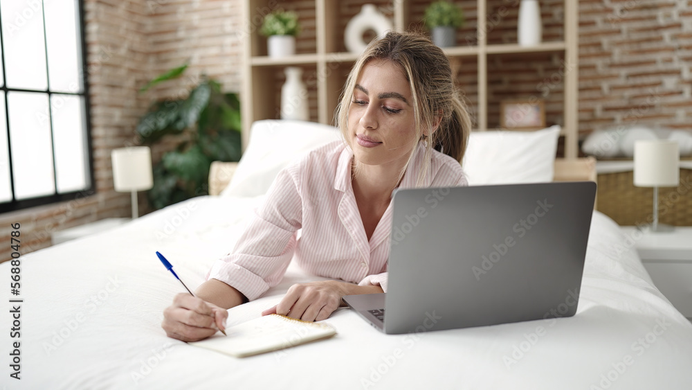 Young blonde woman using laptop writing on notebook at bedroom