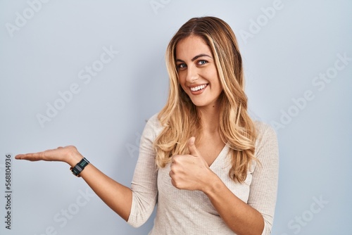 Young blonde woman standing over isolated background showing palm hand and doing ok gesture with thumbs up, smiling happy and cheerful