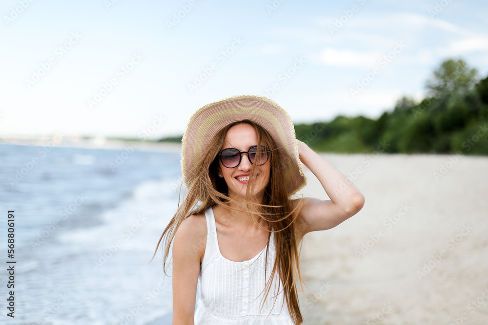 Happy smiling woman in free happiness bliss on ocean beach standing and posing with hat and sunglasses. Portrait of a female model in white summer dress enjoying nature 