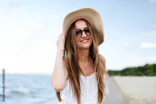 Happy smiling woman in free happiness bliss on ocean beach standing and posing with hat and sunglasses. Portrait of a female model in white summer dress enjoying nature 