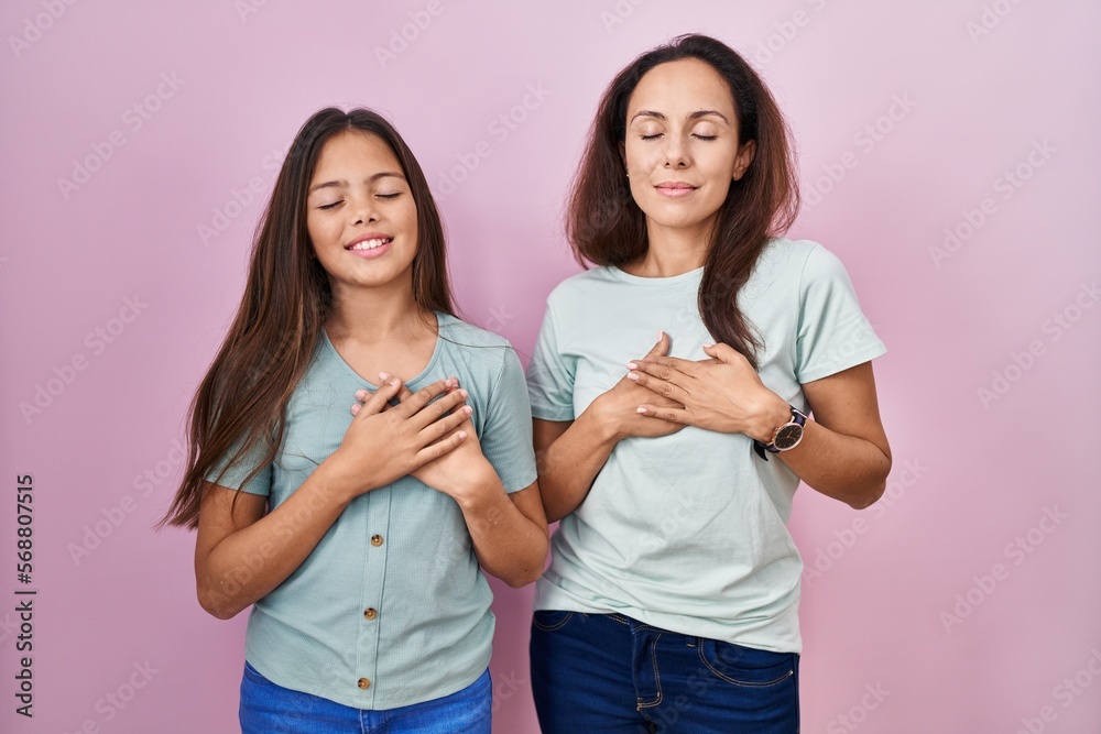 Young mother and daughter standing over pink background smiling with hands on chest with closed eyes and grateful gesture on face. health concept. Young mother and daughter standing over pink background smiling with hands on chest with closed eyes and grateful gesture on face. health concept.