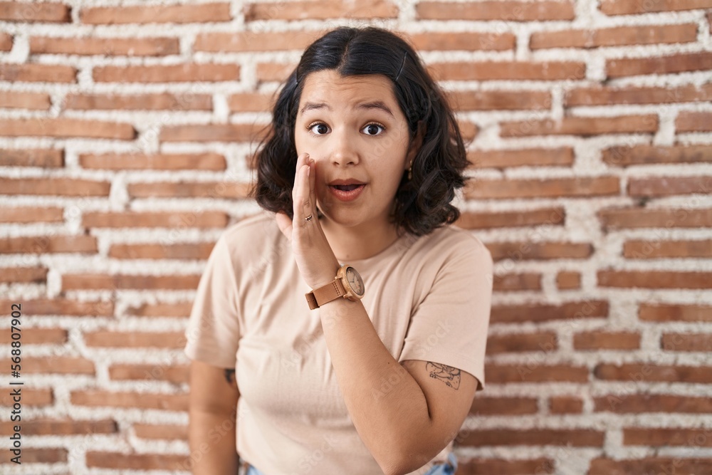 Young hispanic woman standing over bricks wall hand on mouth telling secret rumor, whispering malicious talk conversation Young hispanic woman standing over bricks wall hand on mouth telling secret rumor, whispering malicious talk conversation