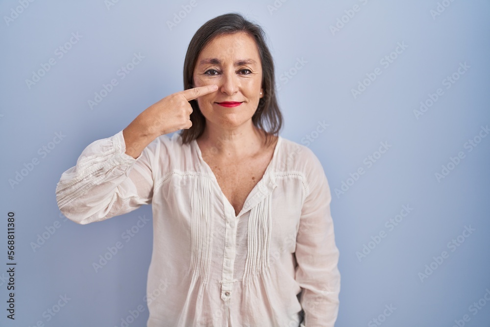 Middle age hispanic woman standing over blue background pointing with hand finger to face and nose, smiling cheerful. beauty concept Middle age hispanic woman standing over blue background pointing with hand finger to face and nose, smiling cheerful. beauty concept