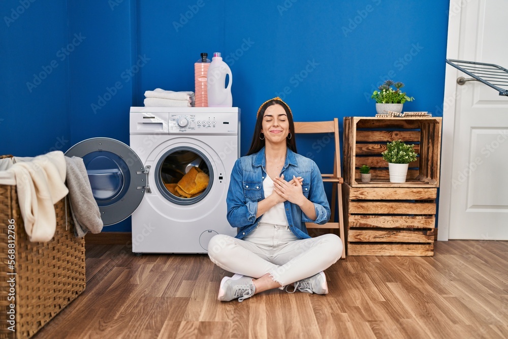 Hispanic woman doing laundry sitting on the floor smiling with hands on chest with closed eyes and grateful gesture on face. health concept. Hispanic woman doing laundry sitting on the floor smiling with hands on chest with closed eyes and grateful gesture on face. health concept.