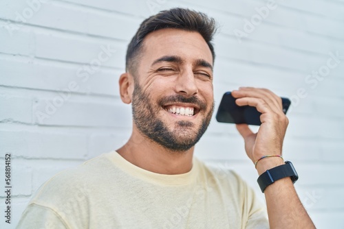 Young hispanic man smiling confident listening audio message by the smartphone over isolated white background