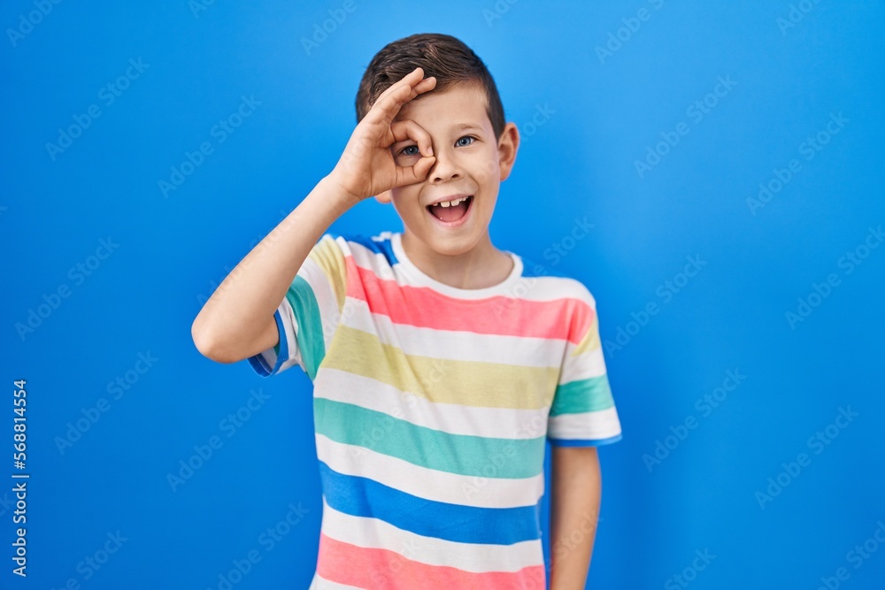 Young caucasian kid standing over blue background doing ok gesture with hand smiling, eye looking through fingers with happy face. Young caucasian kid standing over blue background doing ok gesture with hand smiling, eye looking through fingers with happy face.