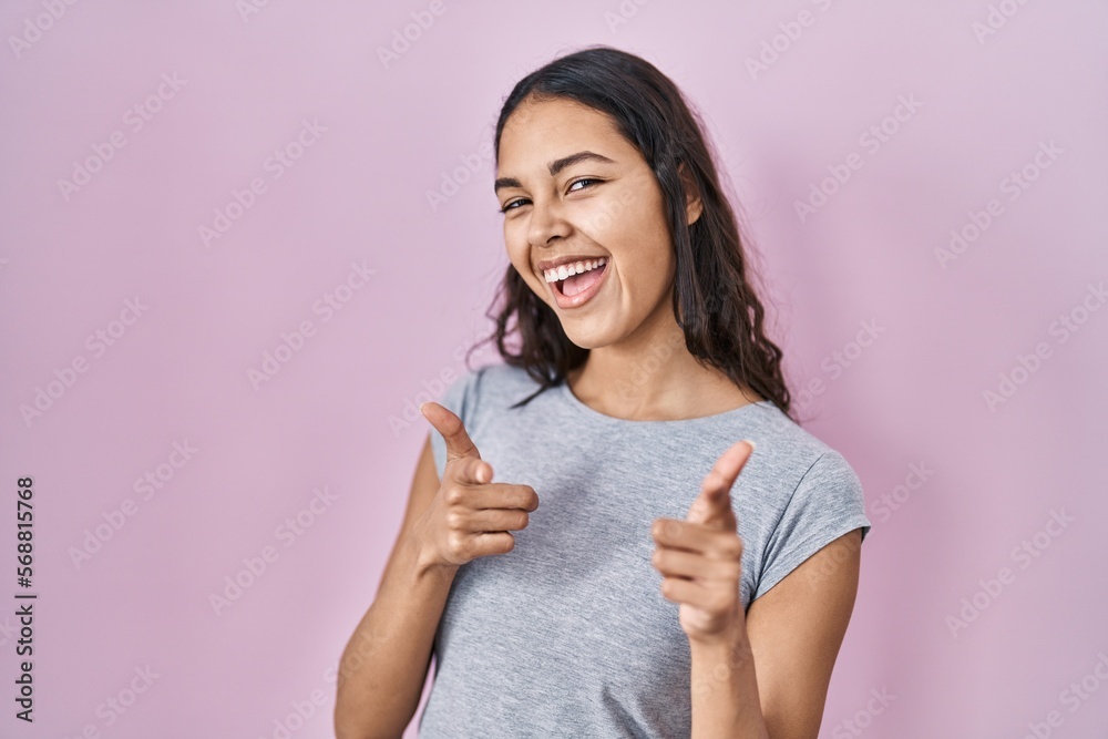 Young brazilian woman wearing casual t shirt over pink background pointing fingers to camera with happy and funny face. good energy and vibes. Young brazilian woman wearing casual t shirt over pink background pointing fingers to camera with happy and funny face. good energy and vibes.