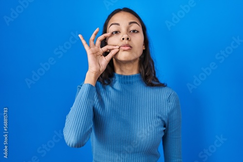 Young brazilian woman standing over blue isolated background mouth and lips shut as zip with fingers. secret and silent, taboo talking