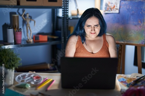 Young modern girl with blue hair sitting at art studio with laptop at night with hand on stomach because nausea, painful disease feeling unwell. ache concept.