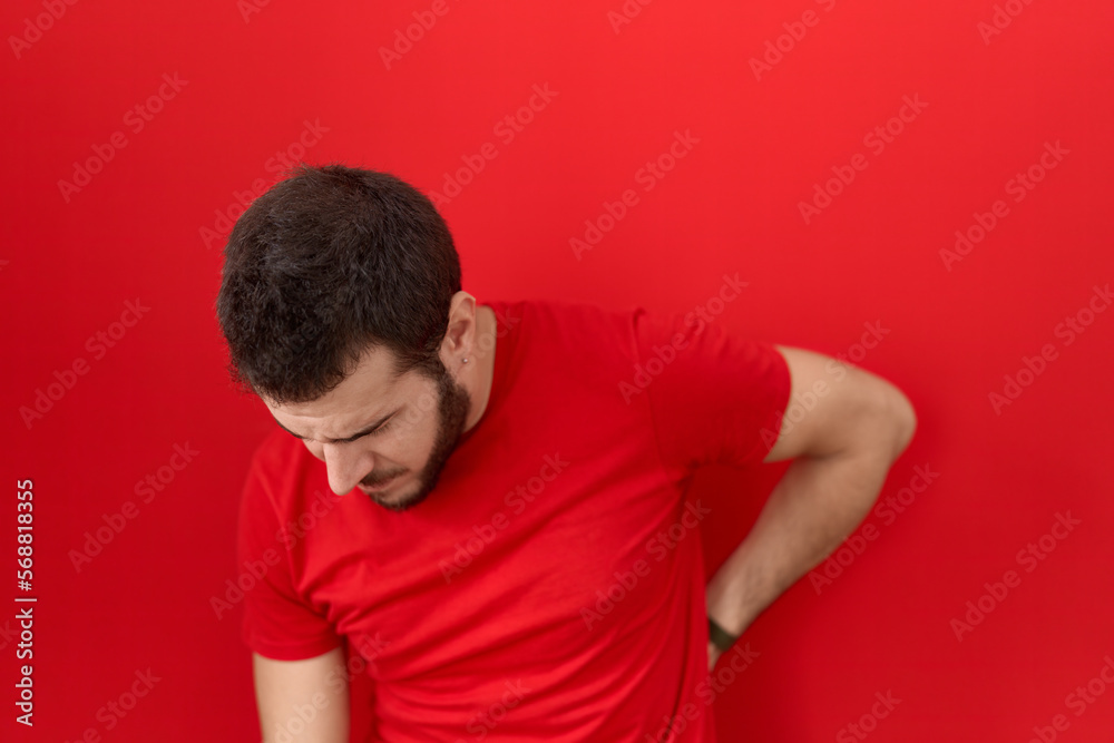 Young hispanic man wearing casual red t shirt suffering of backache, touching back with hand, muscular pain Young hispanic man wearing casual red t shirt suffering of backache, touching back with hand, muscular pain