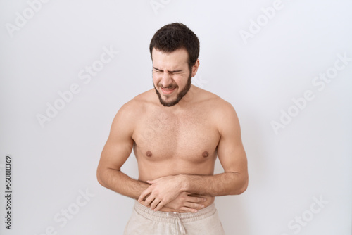Young hispanic man standing shirtless over white background with hand on stomach because nausea, painful disease feeling unwell. ache concept.