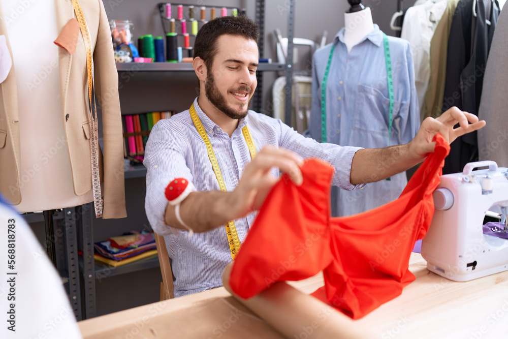 Young hispanic man tailor sitting on table holding t shirt at atelier Young hispanic man tailor sitting on table holding t shirt at atelier