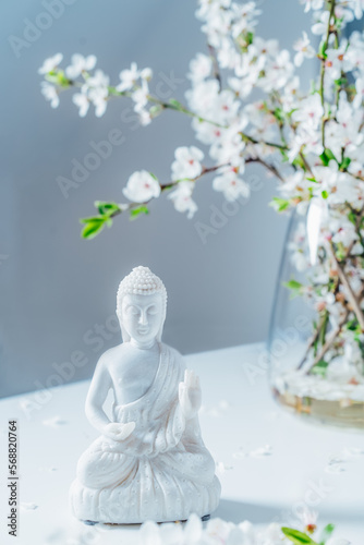 Decorative white Buddha statuette with blooming tree branches in the vase on the light background. Meditation and relaxation ritual. Buddha birthday. Vertical card. Selective focus