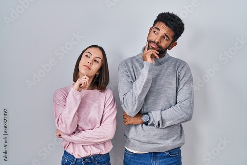 Young hispanic couple standing together with hand on chin thinking about question, pensive expression. smiling with thoughtful face. doubt concept.