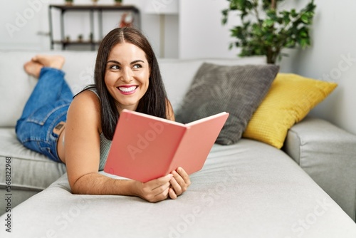 Young beautiful hispanic woman reading book lying on sofa at home