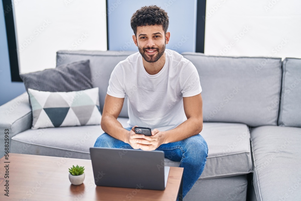 Young arab man using smartphone and laptop sitting on sofa at home Young arab man using smartphone and laptop sitting on sofa at home