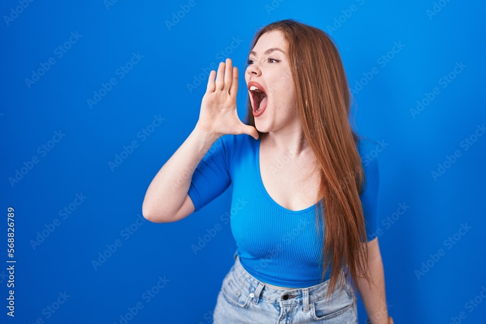 Redhead woman standing over blue background shouting and screaming loud to side with hand on mouth. communication concept. Redhead woman standing over blue background shouting and screaming loud to side with hand on mouth. communication concept.