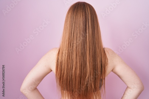 Redhead woman wearing lingerie over pink background standing backwards looking away with arms on body