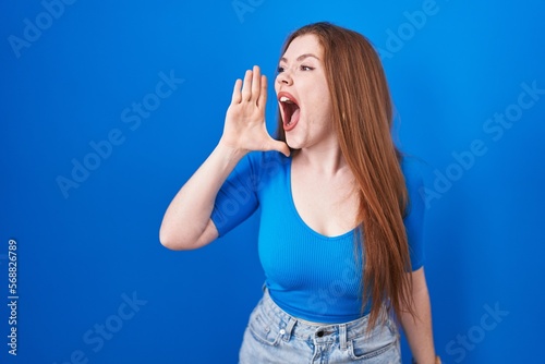 Redhead woman standing over blue background shouting and screaming loud to side with hand on mouth. communication concept.