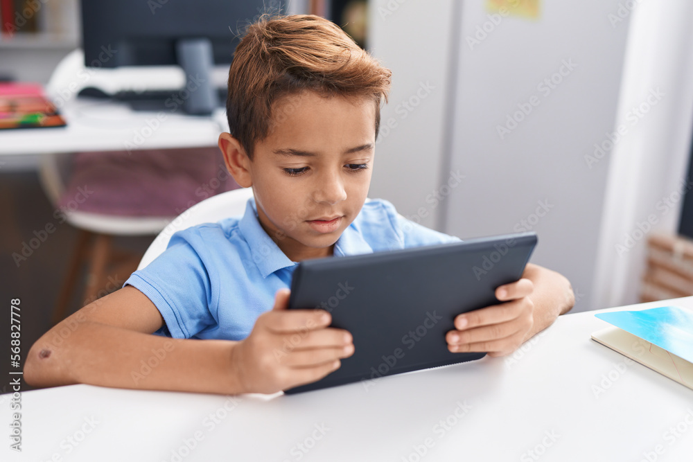 Adorable hispanic boy student using touchpad studying at classroom Adorable hispanic boy student using touchpad studying at classroom