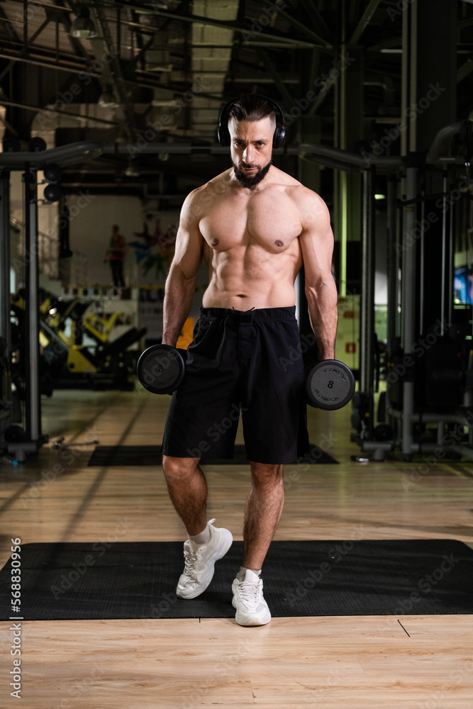 Fototapeta premium A brutal muscular bearded man with a beautiful abs in headphones holds a dumbbell in the gym. Active lifestyle, sports