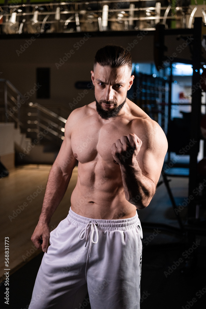 A handsome bearded male athlete with a muscular body poses in the gym in white pants A handsome bearded male athlete with a muscular body poses in the gym in white pants