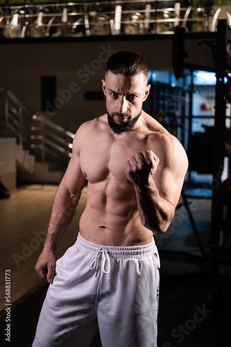 A handsome bearded male athlete with a muscular body poses in the gym in white pants