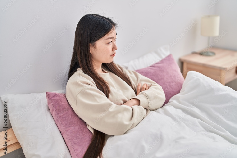Chinese woman sitting on bed with arms crossed gesture and serious expression at bedroom Chinese woman sitting on bed with arms crossed gesture and serious expression at bedroom