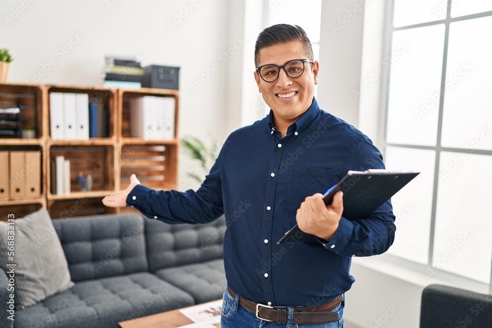 Fototapeta premium Young latin man business worker holding clipboard doing welcome gesture at office