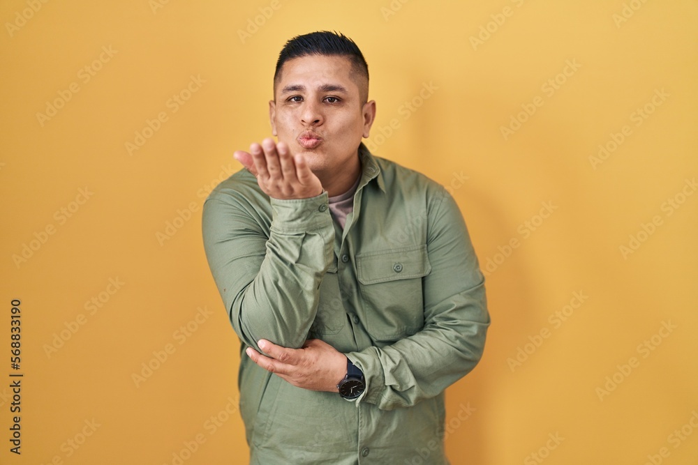 Hispanic young man standing over yellow background looking at the camera blowing a kiss with hand on air being lovely and sexy. love expression. Hispanic young man standing over yellow background looking at the camera blowing a kiss with hand on air being lovely and sexy. love expression.