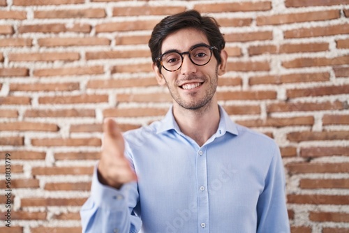 Young hispanic man standing over brick wall background smiling friendly offering handshake as greeting and welcoming. successful business.