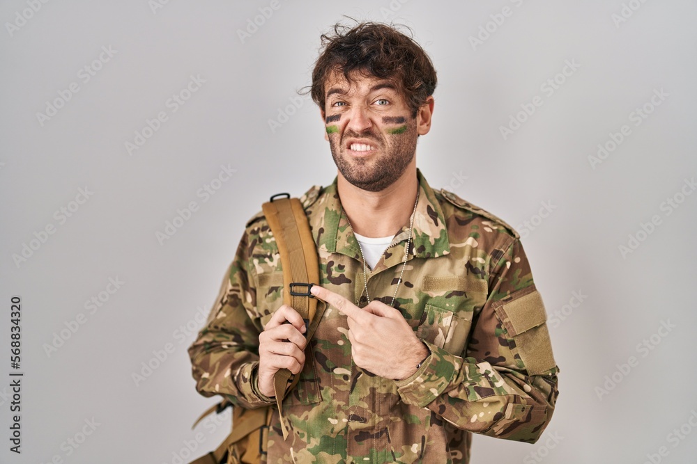 Hispanic young man wearing camouflage army uniform pointing aside worried and nervous with forefinger, concerned and surprised expression