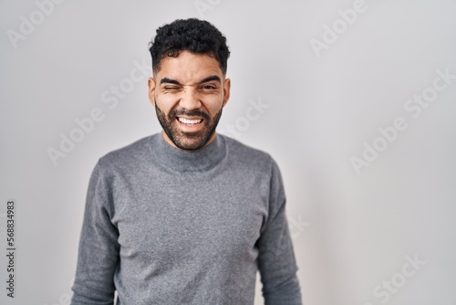 Hispanic man with beard standing over white background winking looking at the camera with sexy expression, cheerful and happy face. photo