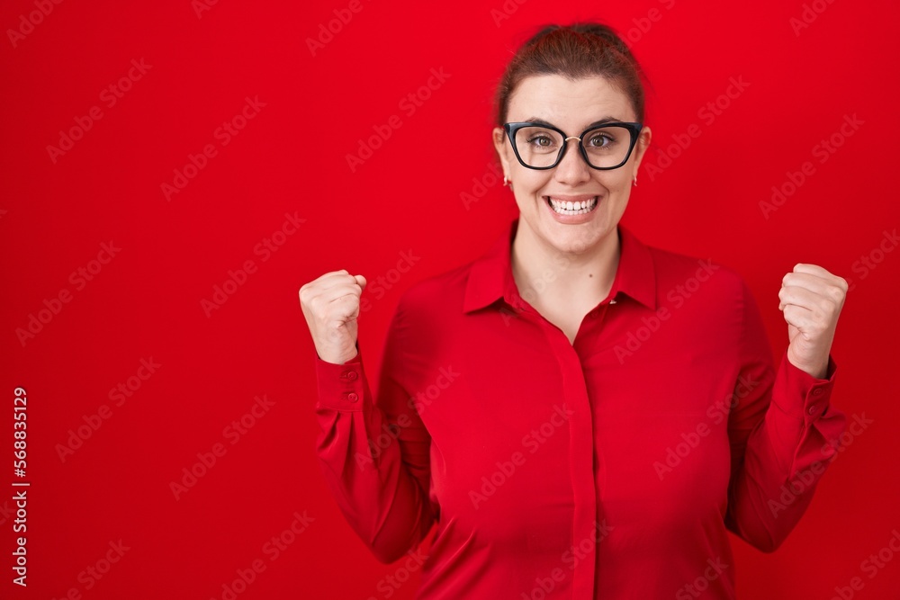 Young hispanic woman with red hair standing over red background celebrating surprised and amazed for success with arms raised and open eyes. winner concept.