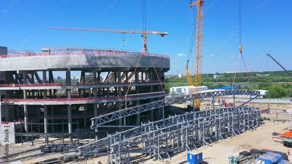 Metal structure near concrete foundation of athletic arena against blue sky. Cranes operate at construction site in city aerial view Metal structure near concrete foundation of athletic arena against blue sky. Cranes operate at construction site in city aerial view
