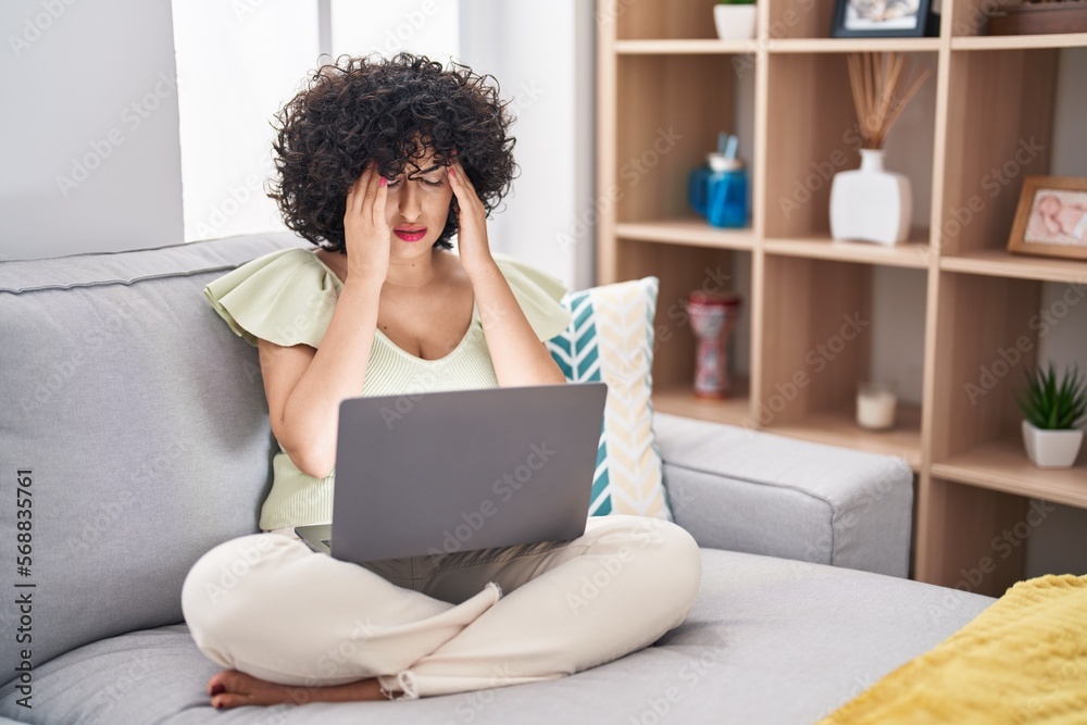 Young brunette woman with curly hair using laptop sitting on the sofa at home with hand on head for pain in head because stress. suffering migraine. Young brunette woman with curly hair using laptop sitting on the sofa at home with hand on head for pain in head because stress. suffering migraine.