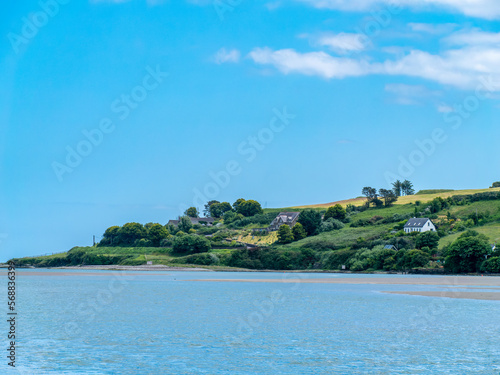 Beautiful clear sky over a calm water surface on a summer day. The picturesque green coast of Ireland. Several buildings on the hill. Seascape.