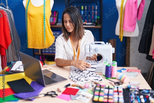 Young hispanic woman tailor using sewing machine and laptop at sewing studio photo