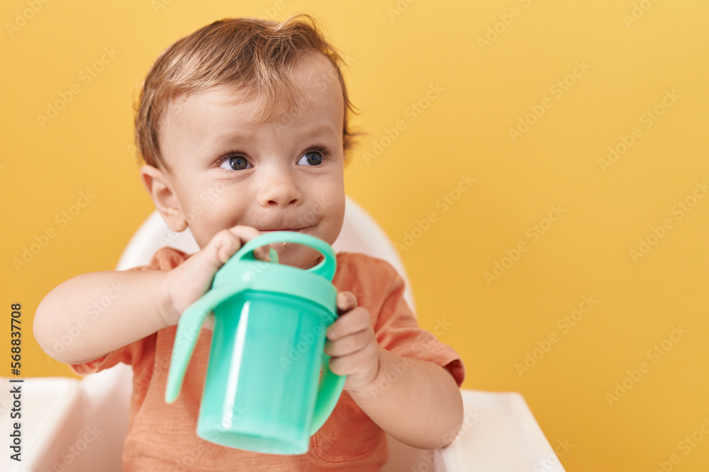 Adorable blond toddler sitting on highchair drinking over isolated yellow background Adorable blond toddler sitting on highchair drinking over isolated yellow background