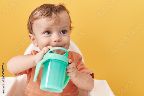 Adorable blond toddler sitting on highchair drinking over isolated yellow background