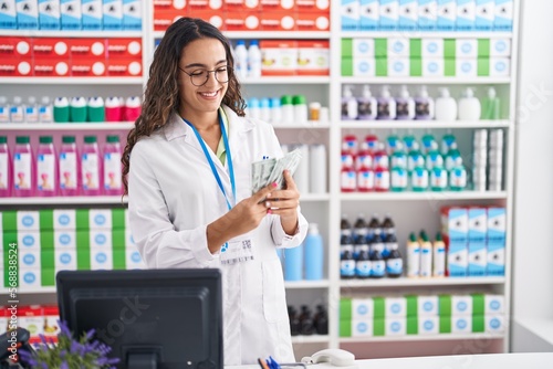Young beautiful hispanic woman pharmacist smiling confident counting dollars at pharmacy