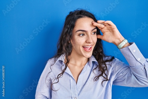 Young brunette woman standing over blue background very happy and smiling looking far away with hand over head. searching concept.
