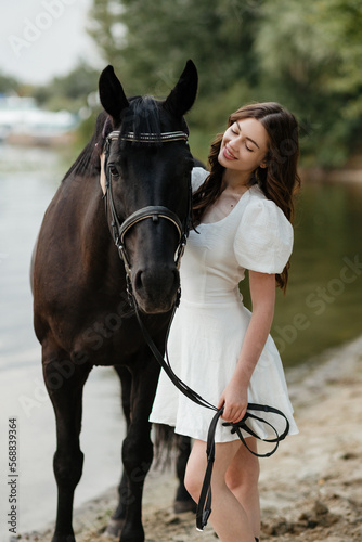 A beautiful girl in a white dress leads a horse along the shore