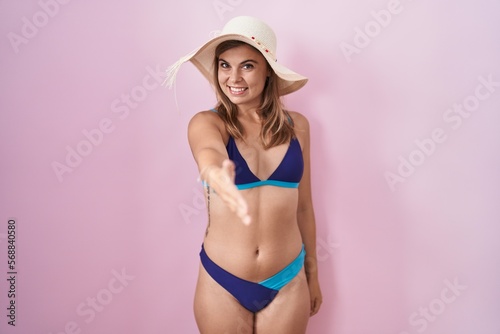 Young hispanic woman wearing bikini over pink background smiling friendly offering handshake as greeting and welcoming. successful business.