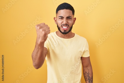 Young hispanic man standing over yellow background angry and mad raising fist frustrated and furious while shouting with anger. rage and aggressive concept.