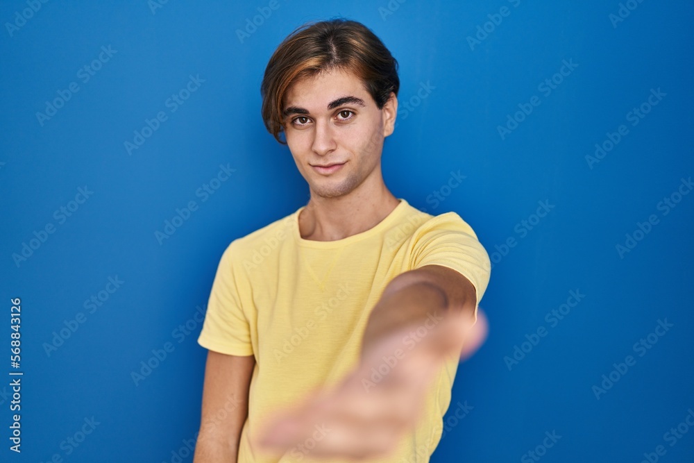 Young man standing over blue background smiling friendly offering handshake as greeting and welcoming. successful business. Young man standing over blue background smiling friendly offering handshake as greeting and welcoming. successful business.